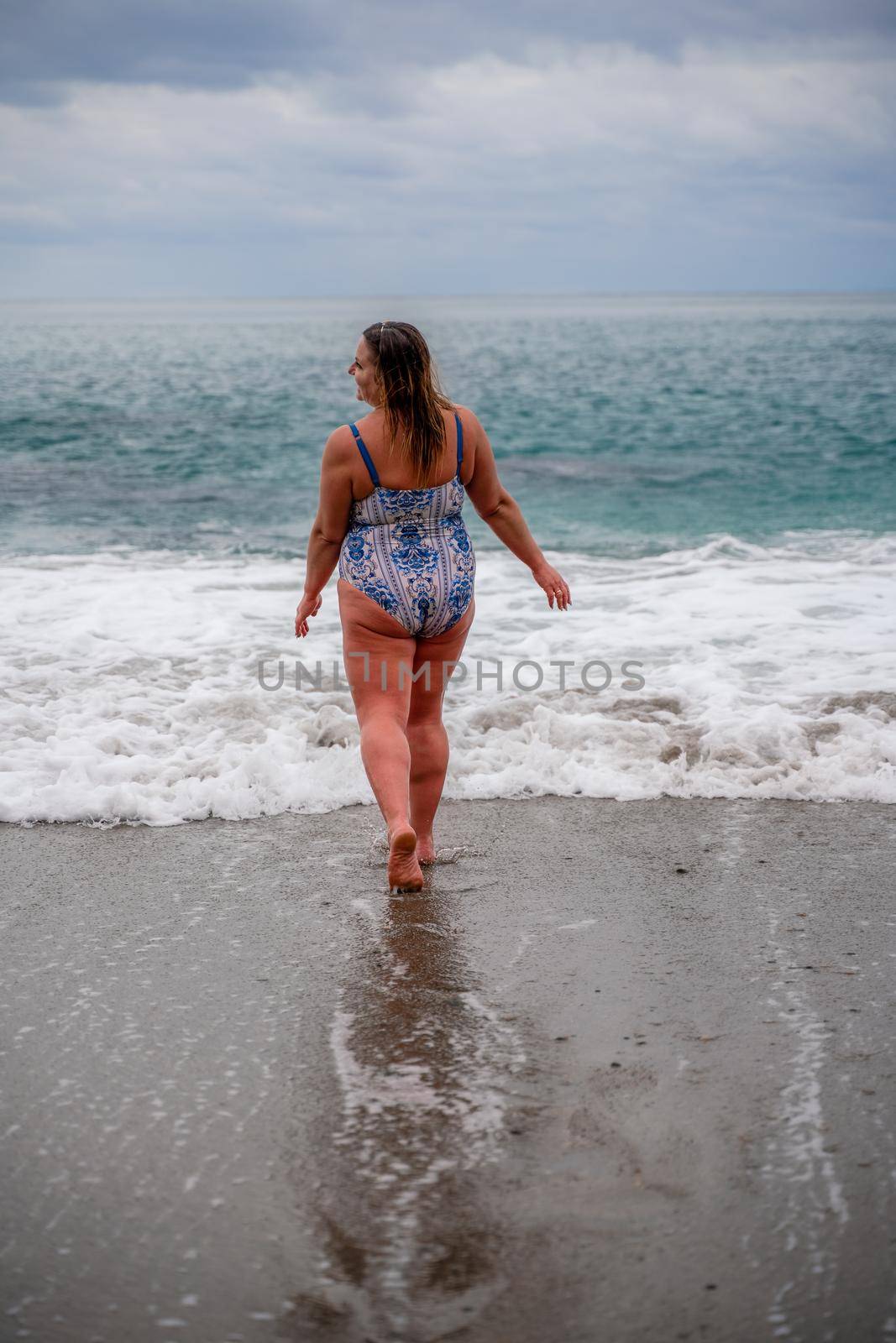 A plump woman in a bathing suit enters the water during the surf. Alone on the beach, Gray sky in the clouds, swimming in winter