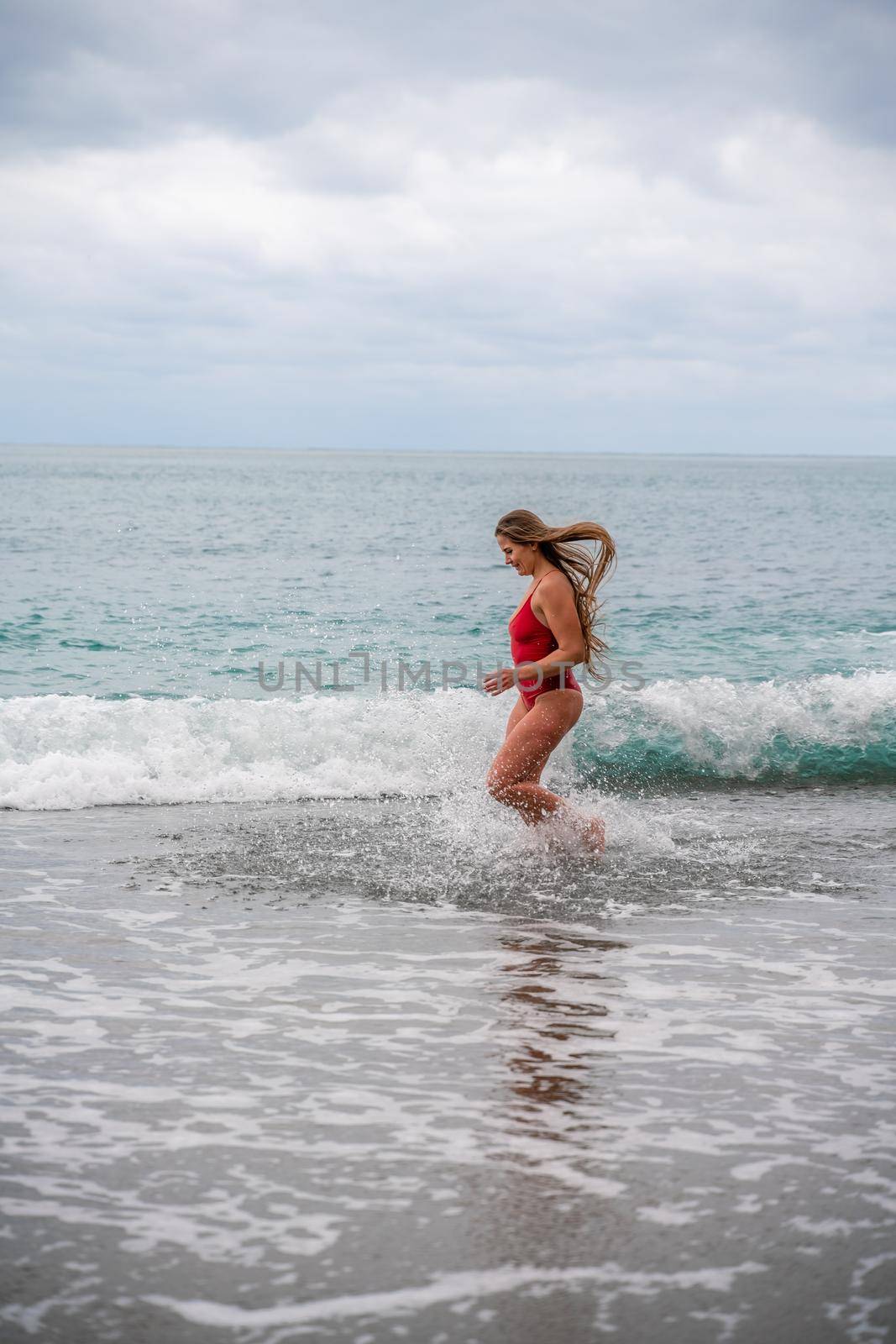 A beautiful and sexy brunette in a red swimsuit on a pebble beach, Running along the shore in the foam of the waves.