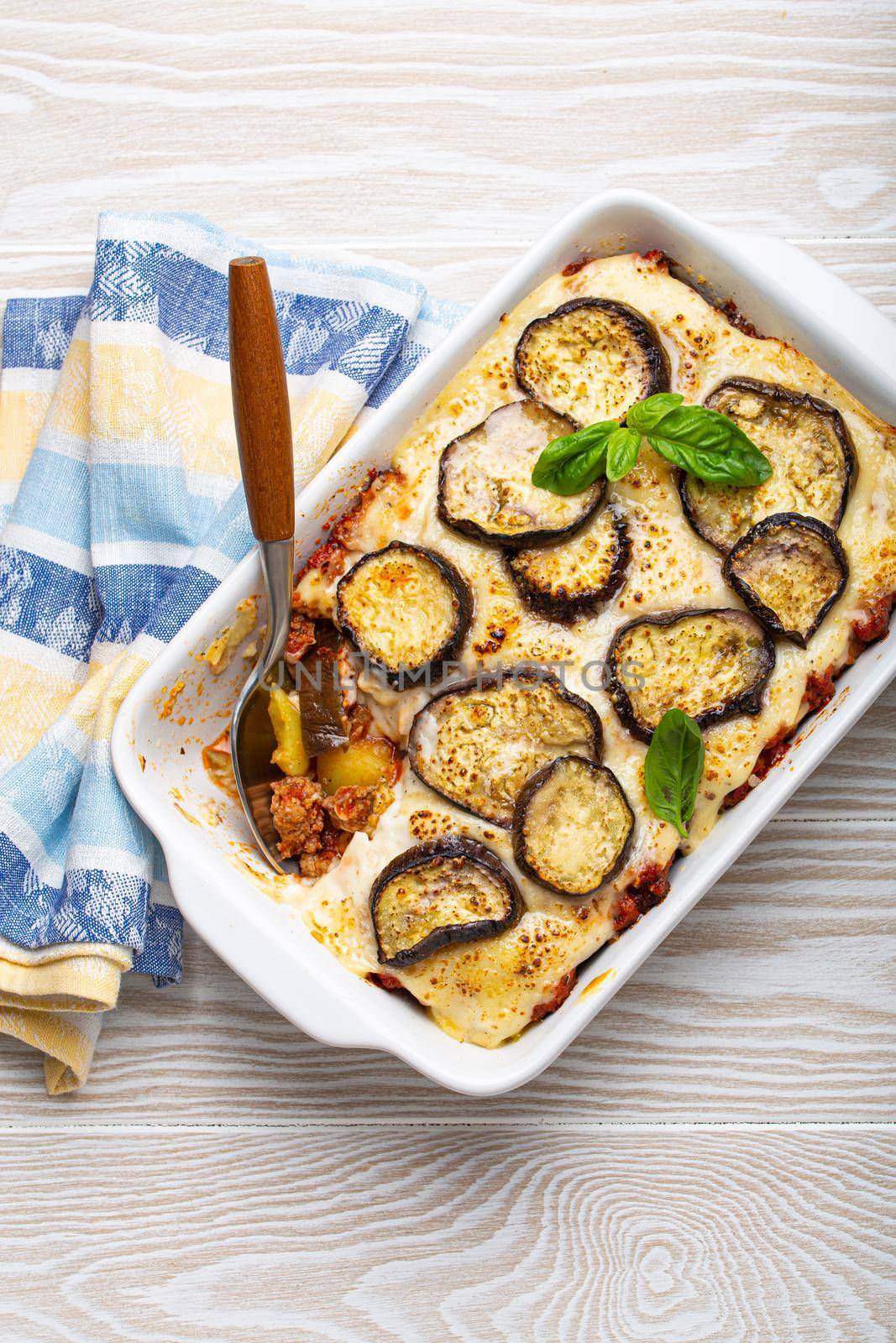 Greek mediterranean dish Moussaka with baked eggplants, ground beef in white ceramic casserole with napkin on wooden white background from above, traditional dish of Greece