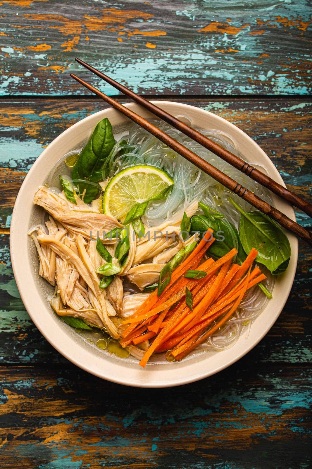 Asian soup with rice noodles, chicken and vegetables in ceramic bowl served with spoon and chopsticks on rustic wooden background from above, Chinese or Thai cuisine