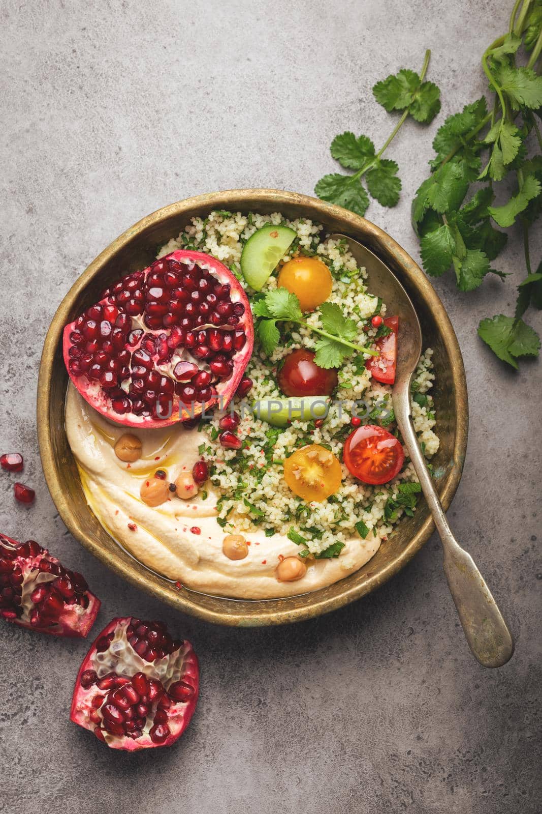 Rustic bowl with couscous salad with vegetables, hummus and fresh cut pomegranate. Middle eastern or Arab style meal with seasonings and fresh cilantro. Healthy Mediterranean dinner, toned image.