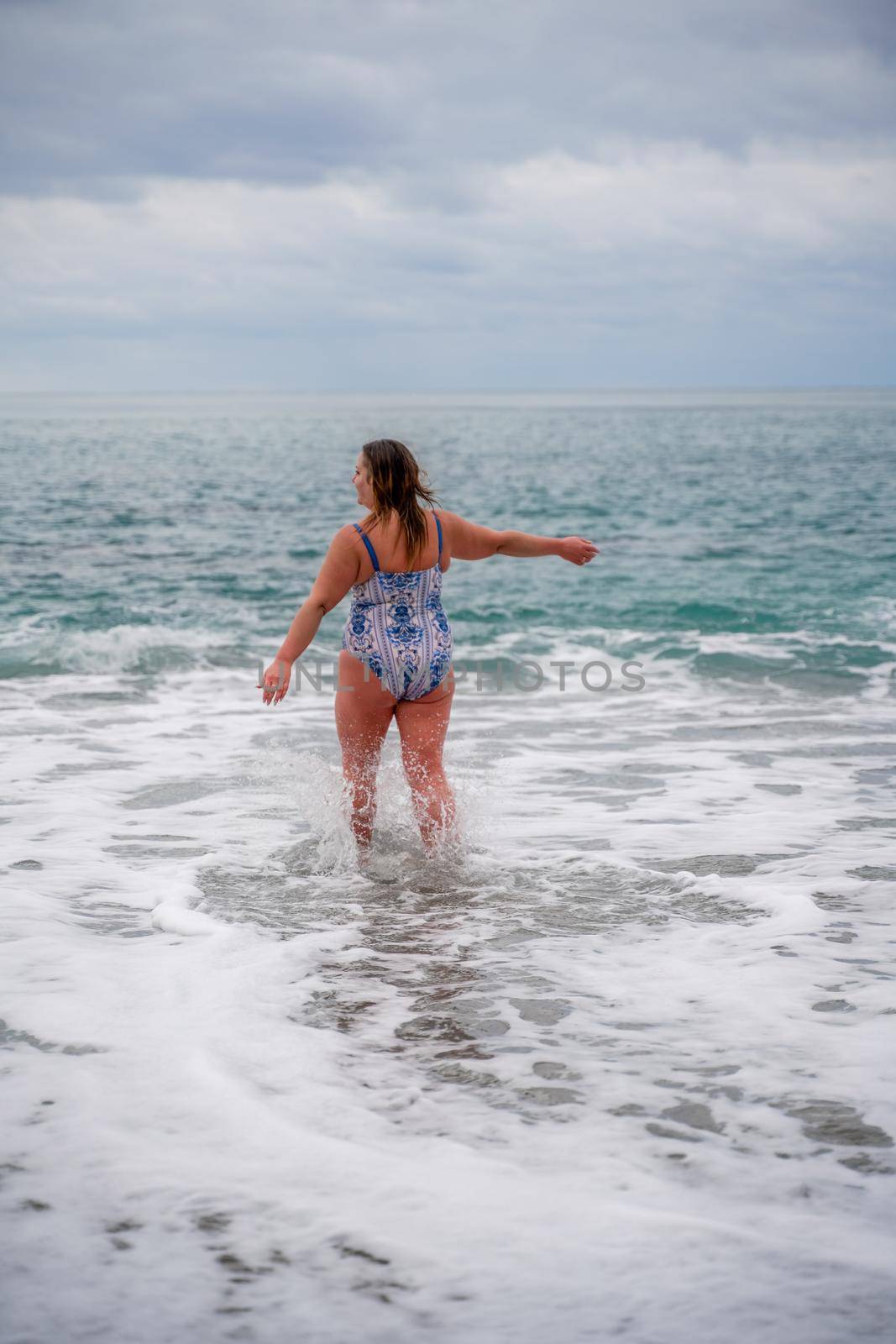 A plump woman in a bathing suit enters the water during the surf. Alone on the beach, Gray sky in the clouds, swimming in winter. by Matiunina