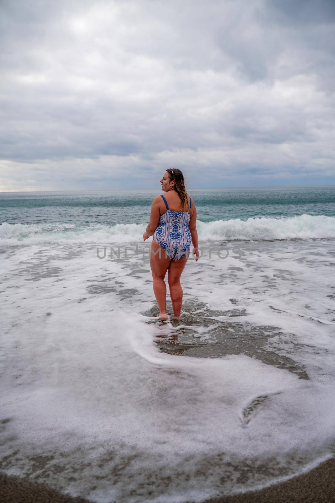 A plump woman in a bathing suit enters the water during the surf. Alone on the beach, Gray sky in the clouds, swimming in winter