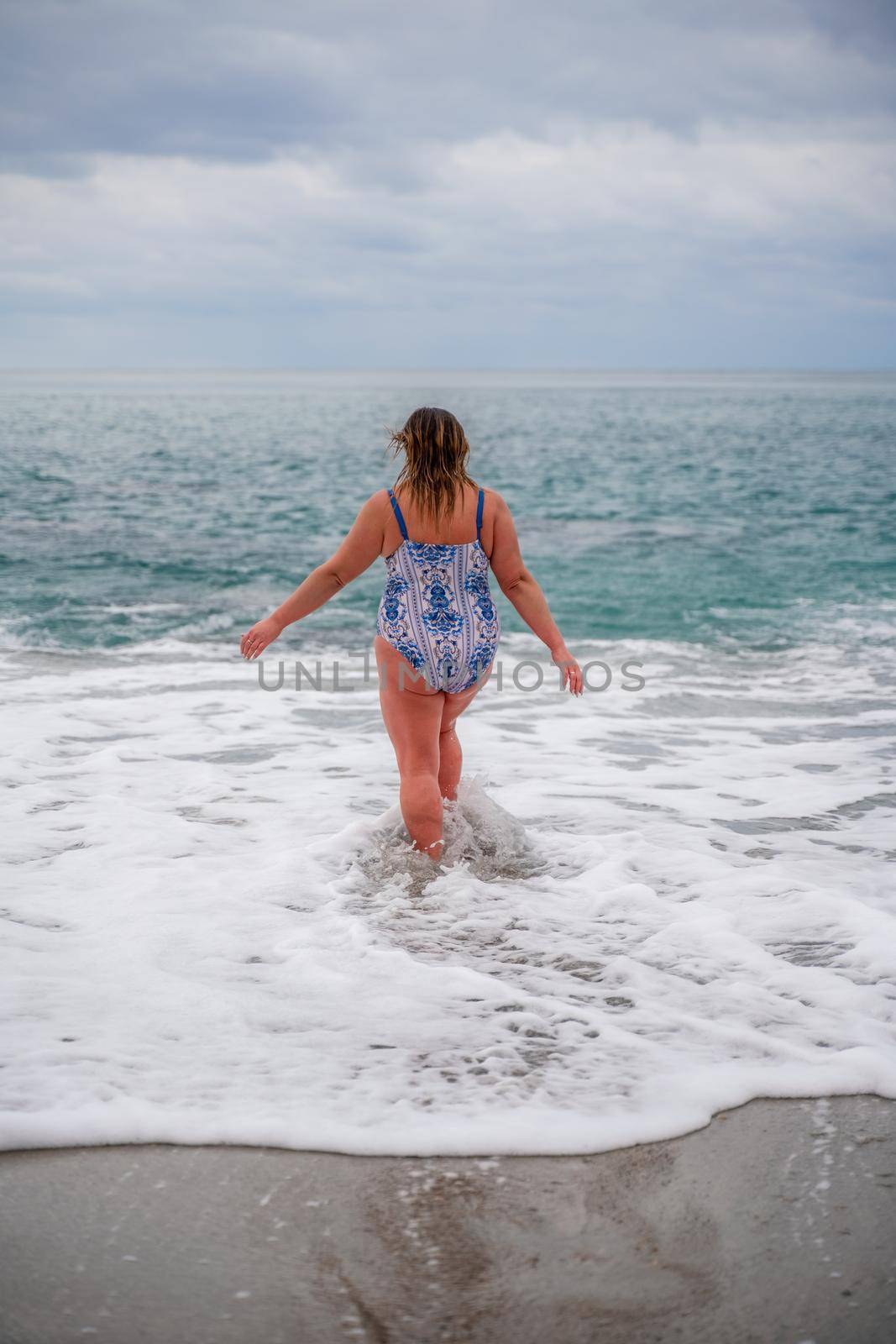 A plump woman in a bathing suit enters the water during the surf. Alone on the beach, Gray sky in the clouds, swimming in winter