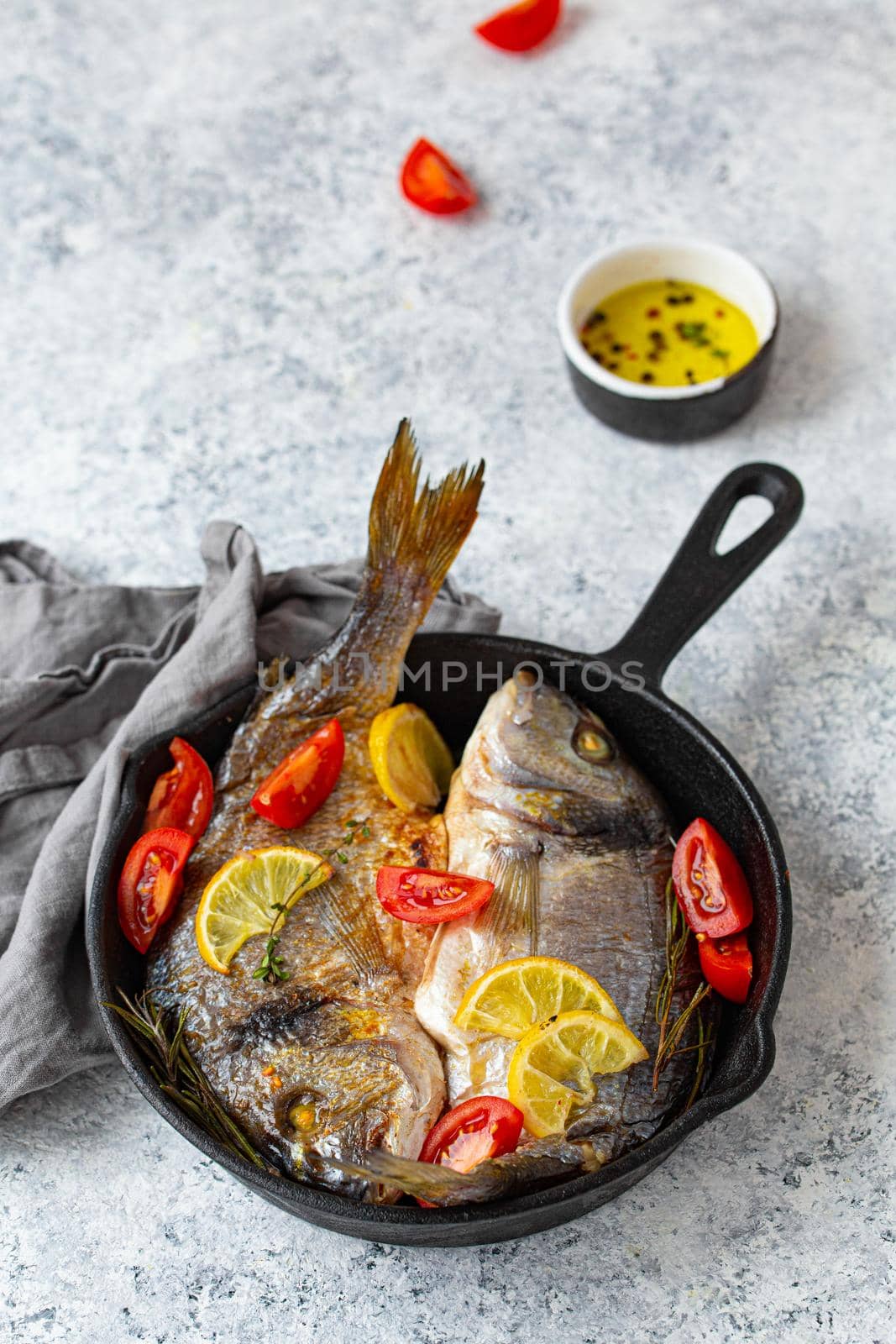 Roasted or grilled fish sea bream or dorado with lemon, herbs and tomatoes in cast iron skillet great for healthy meal or Mediterranean diet on rustic white stone table, angle view