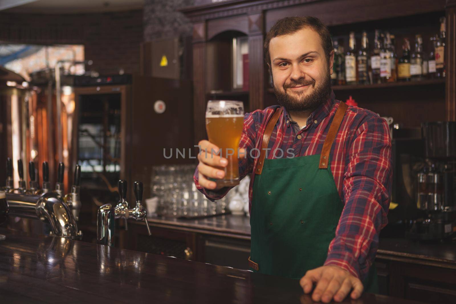 Happy bearded bartender wearing apron, smiling cheerfully to the camera, holding glass of beer, posing behind the counter. Friendly brewer working at his pub, copy space. Recreation concept