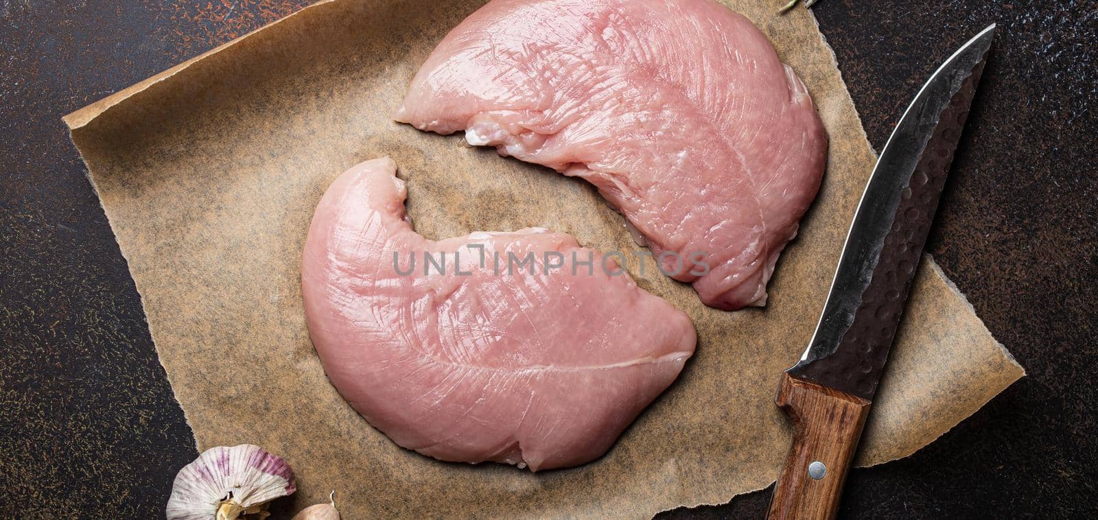 Turkey lean raw fillet on baking paper with rosemary, garlic and spices on dark brown rustic concrete background with knife from above flat lay, healthy diet turkey meat ready to be cooked