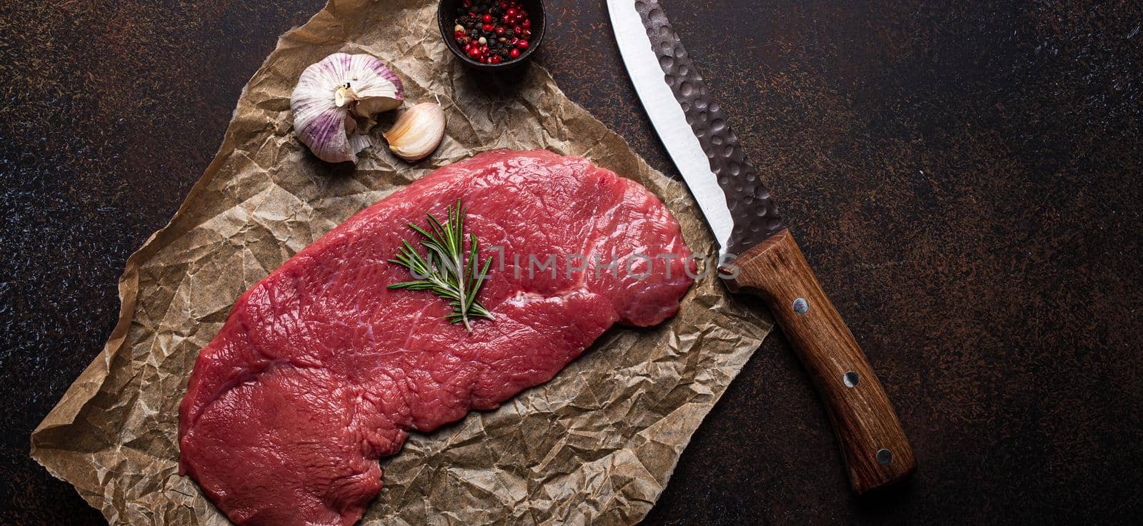 Beef lean raw fillet steak on baking paper with rosemary, garlic and spices on dark brown rustic concrete background with knife from above flat lay, diet beef meat steak ready to be cooked, copy space