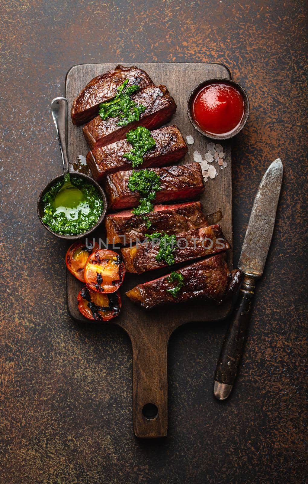 Grilled or fried and sliced marbled meat steak with fork, tomatoes as a side dish and different sauces on wooden cutting board, top view, close-up, stone rustic background. Beef meat steak concept