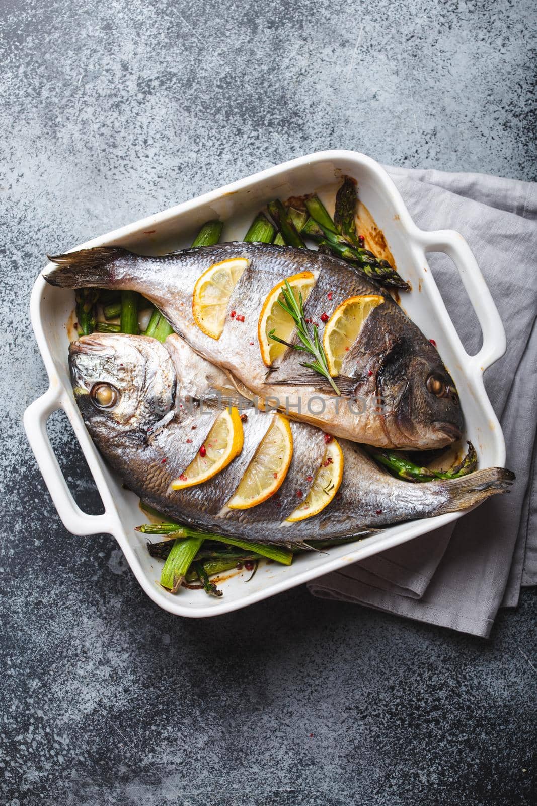 Baked fish dorado with green asparagus in white ceramic baking pan on gray rustic concrete background, top view. Healthy dinner with fish concept, dieting and clean eating .