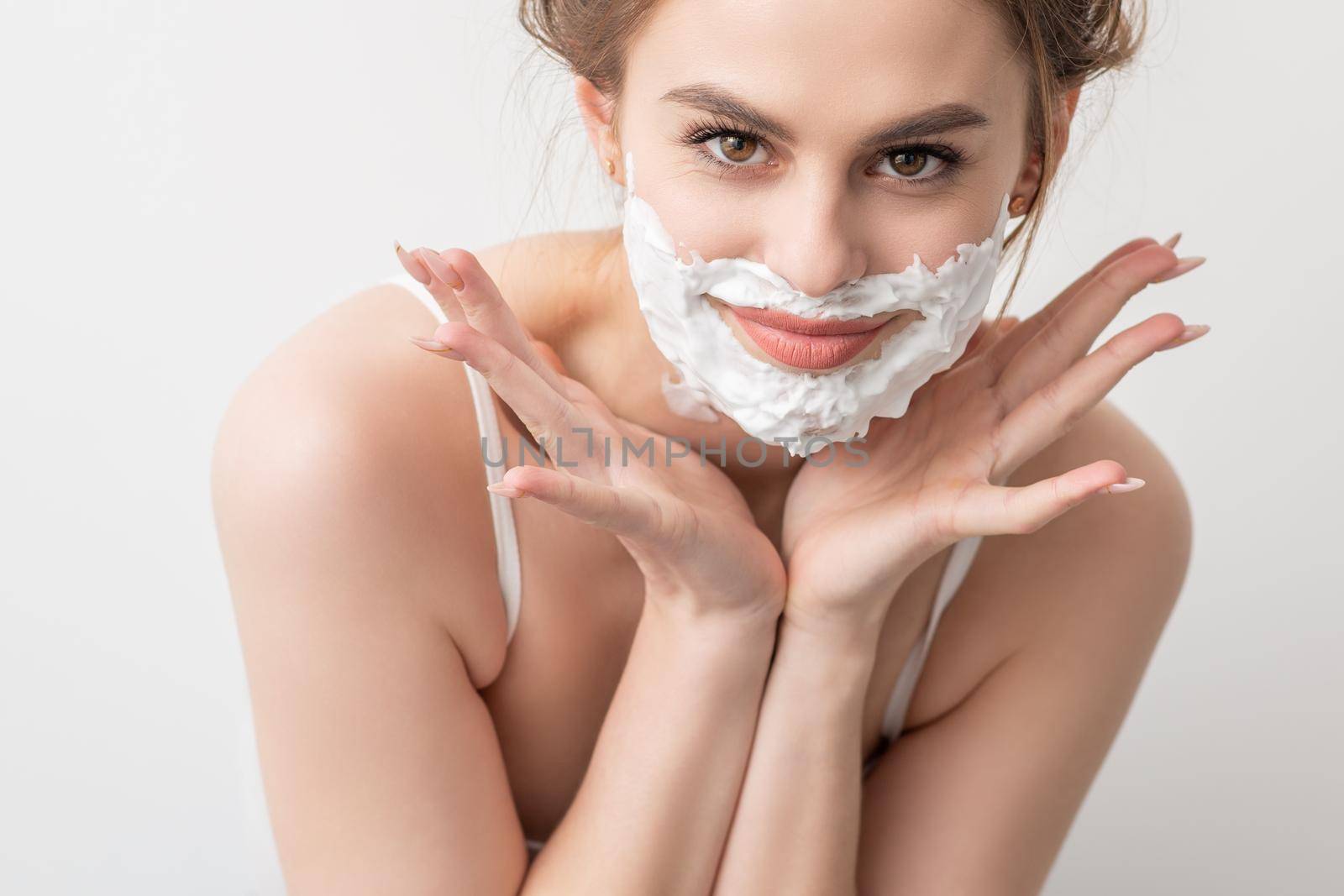 Portrait of a beautiful young smiling caucasian woman with shaving foam on face poses on white background