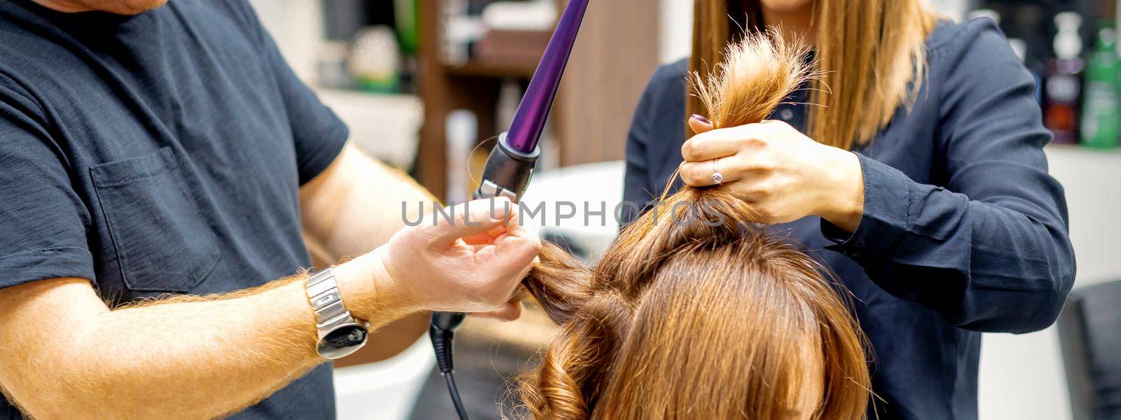 Two hairstylists using curling iron on customers long brown hair in a beauty salon