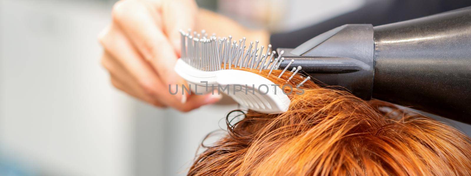 Closeup of master's hand with blow-drying and hairbrush blowing female red hair in a salon. by okskukuruza