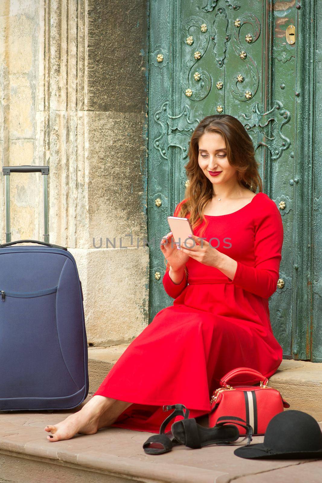 Beautiful young caucasian woman sitting on the stairs by the door with travel suitcase and smartphone wearing long red dress