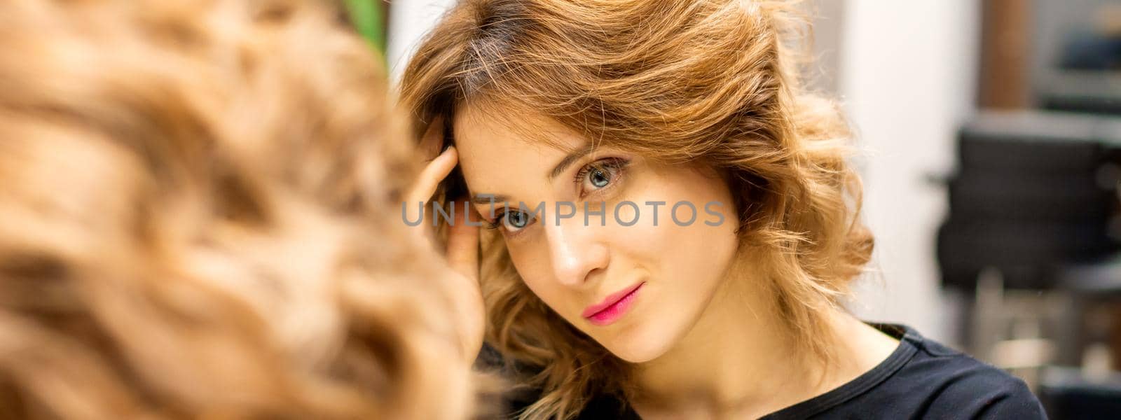 The beautiful young caucasian woman looks at her reflection in the mirror checking hairstyle and makeup in a beauty salon