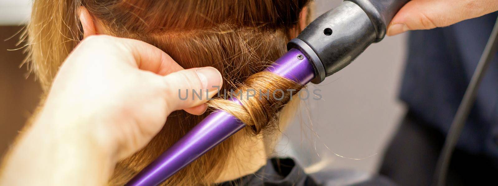 Close up of hairstylist's hands using a curling iron for hair curls in a beauty salon. by okskukuruza