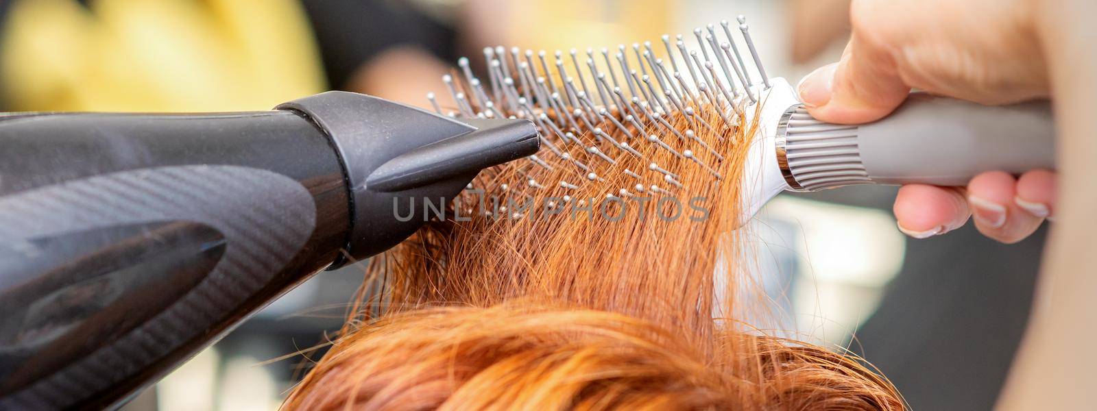 Closeup of master's hand with blow-drying and hairbrush blowing female red hair in a salon. by okskukuruza