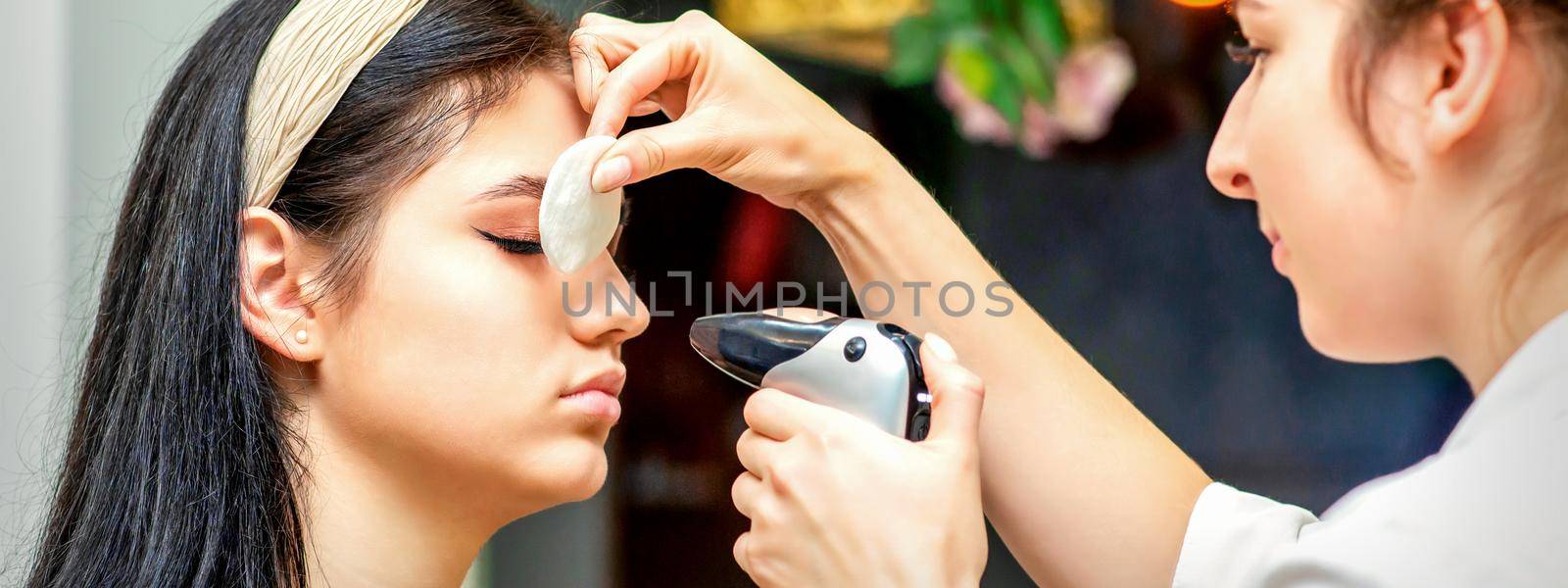 Side view of a make-up artist using aerograph making an airbrush makeup foundation on a female face in a beauty salon. by okskukuruza