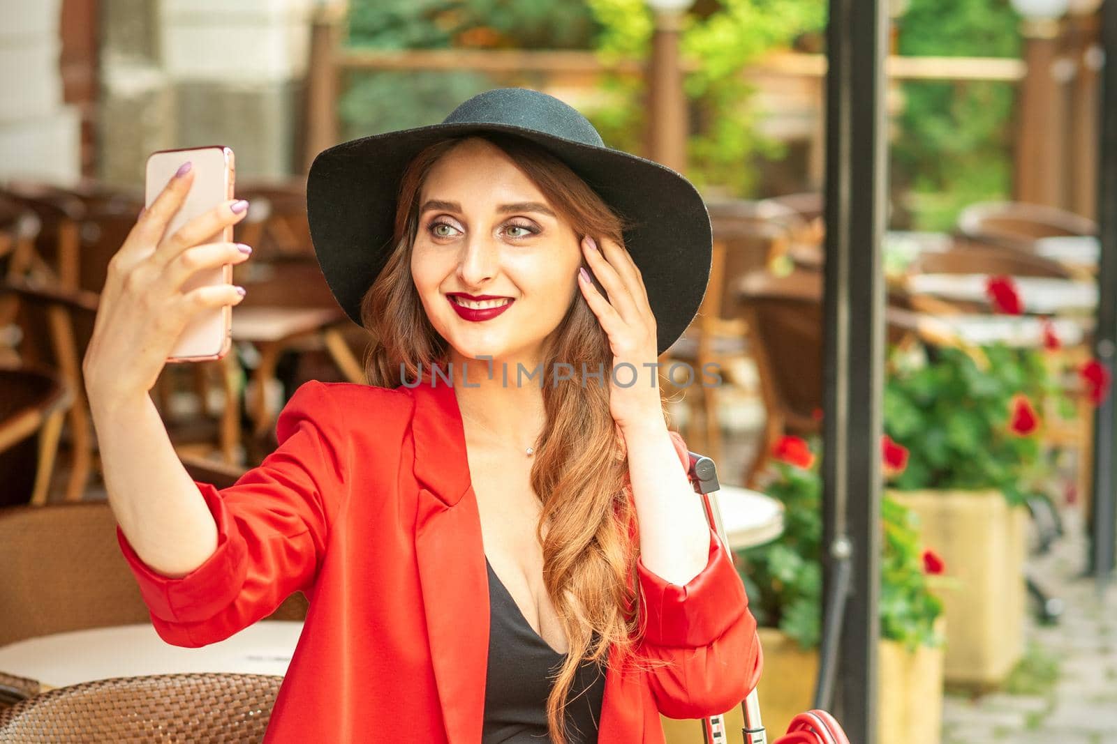 Tourist young caucasian woman in a red jacket and black hat with suitcase takes a selfie on a city street