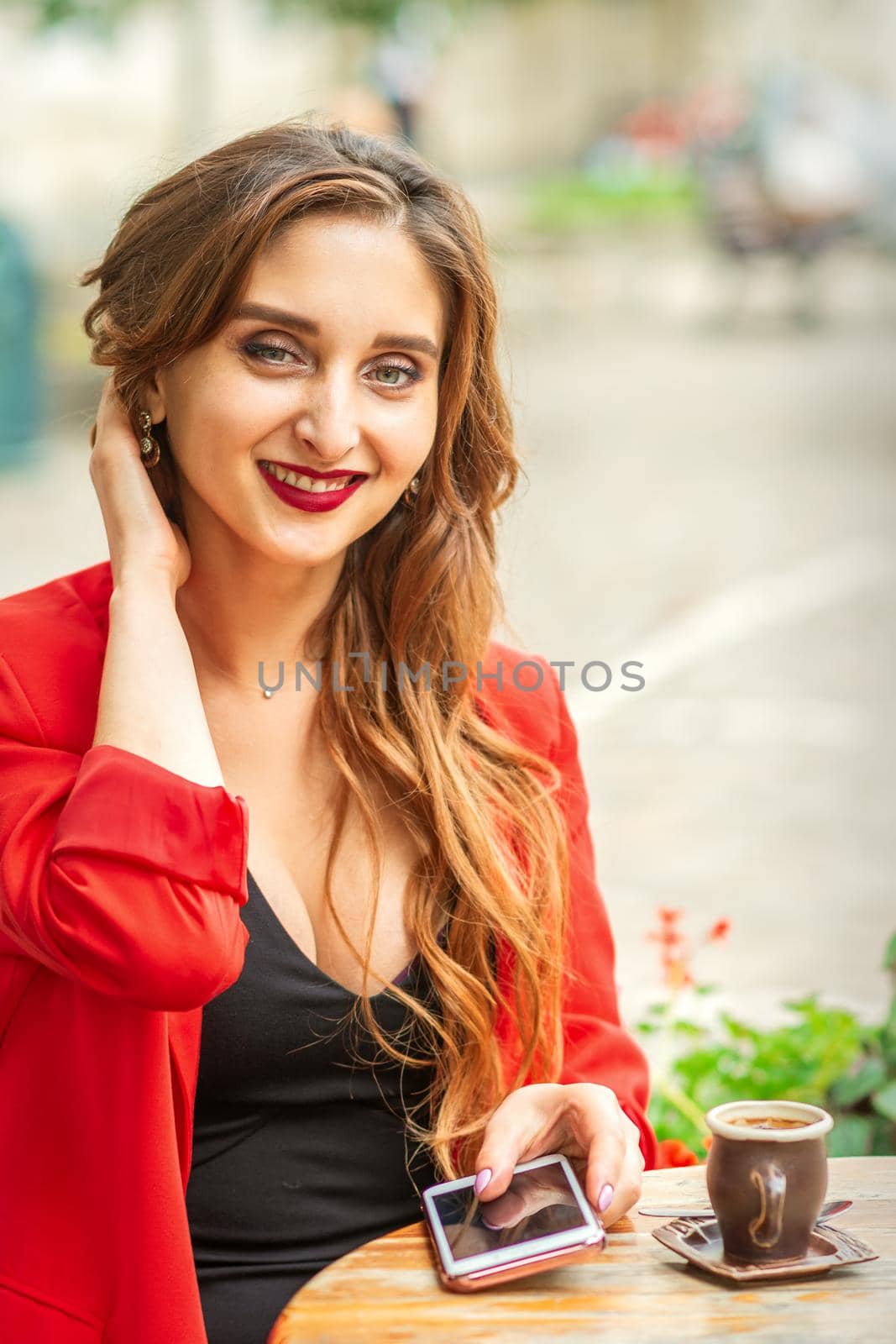 Portrait of a young white woman sitting at the table with a smartphone and looking at camera outdoors