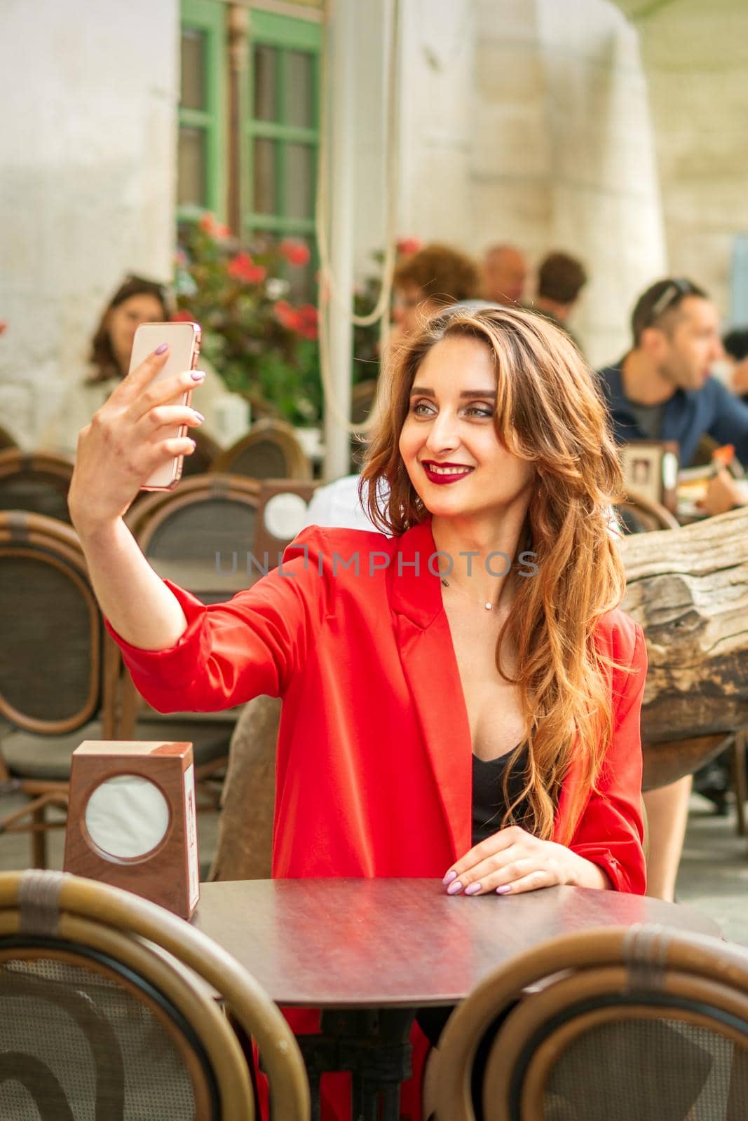 Tourist young caucasian woman in a red jacket with suitcase takes a selfie at the table in cafe outdoors