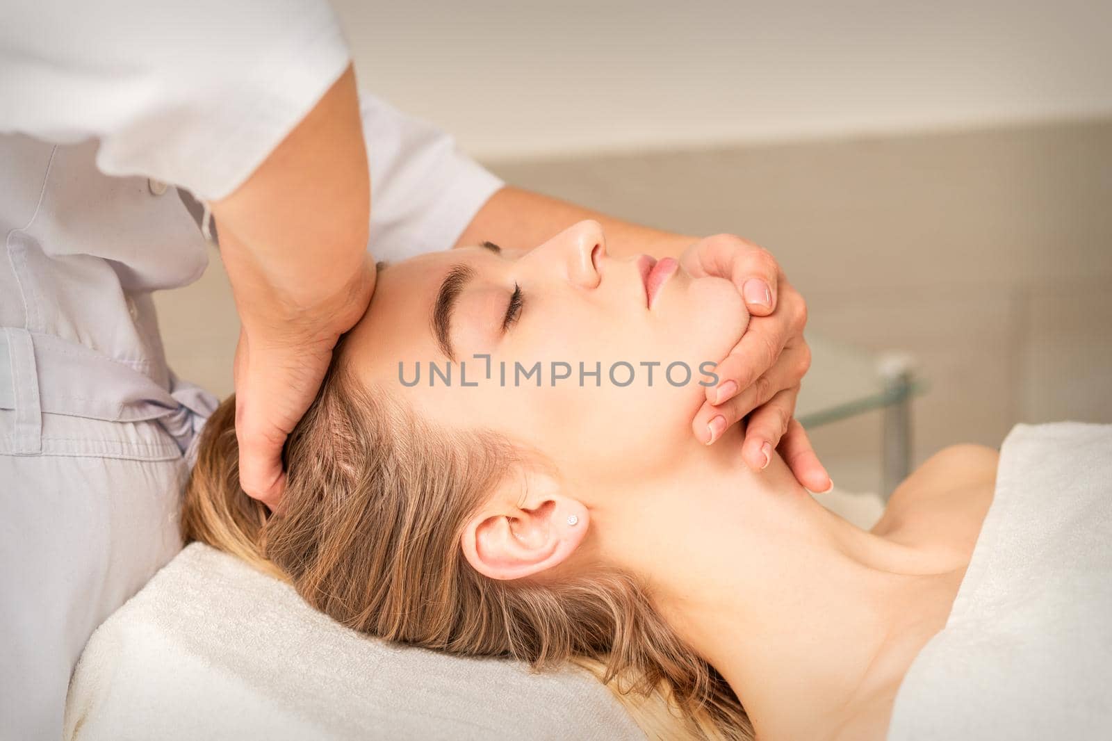 Cosmetologist hands doing facial massage on forehead and chin of young female at spa salon