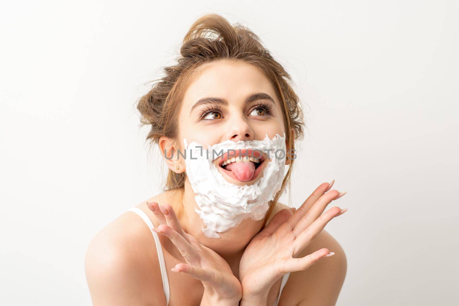 Portrait of a beautiful young smiling caucasian woman posing with shaving foam on the face stuck out her tongue on white background