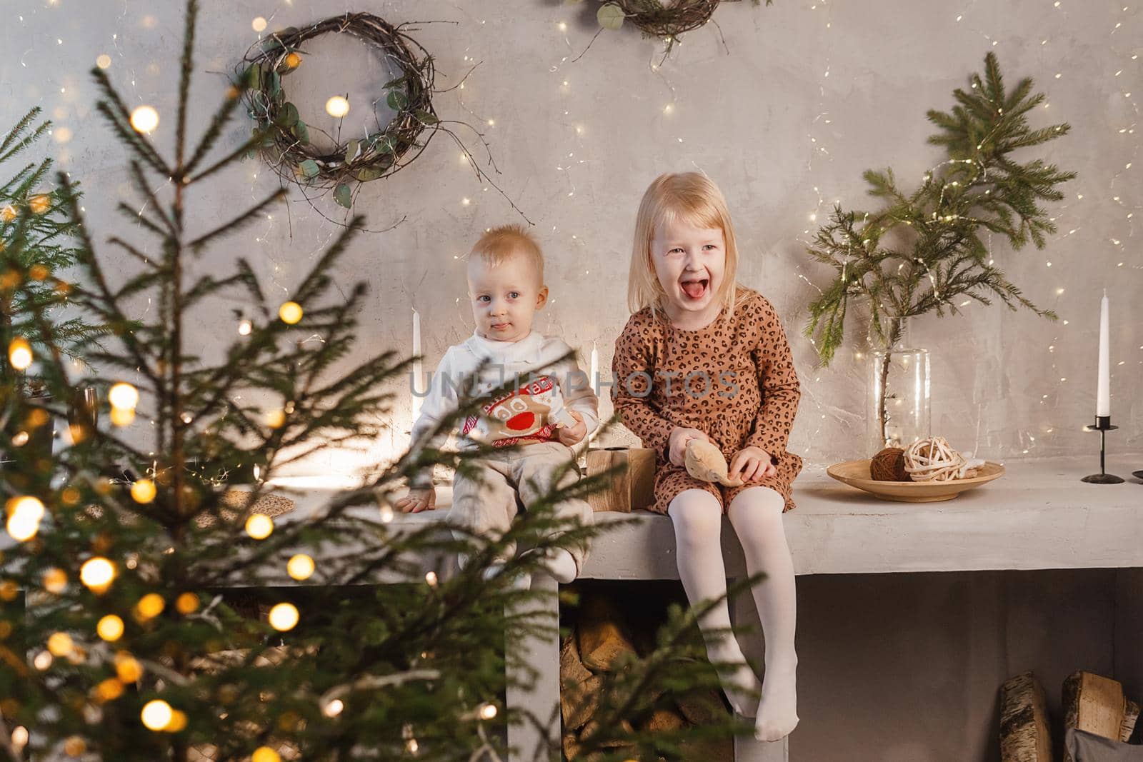 Little brother and sister play on Christmas eve in a beautiful house decorated for the New Year holidays. Children are playing with a Christmas gift. Scandinavian-style interior with live fir trees and a wooden staircase.
