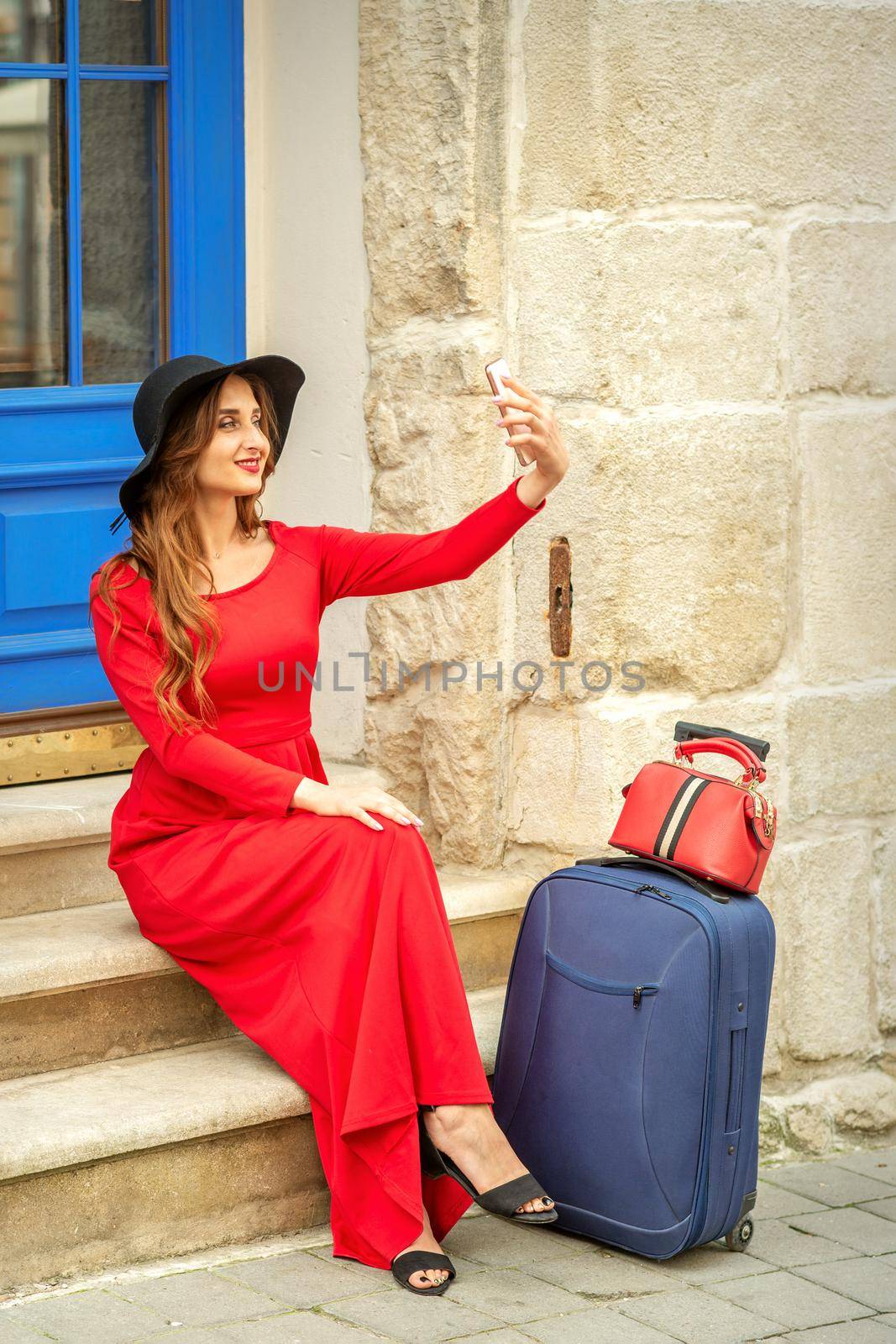 Tourist young caucasian woman in a red long dress and black hat with suitcase takes a selfie sitting on the stairs at the door outdoors