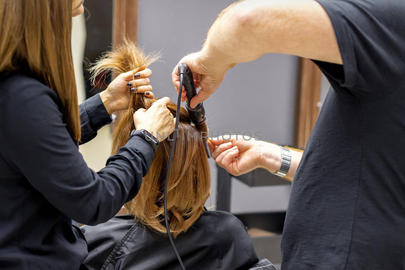 The rear view of two hairdressers are curling hair for a young woman with electric hair iron in a beauty salon