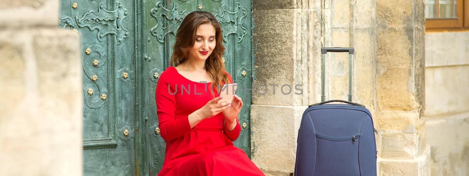 Beautiful young caucasian woman sitting on the stairs by the door with travel suitcase and smartphone wearing long red dress