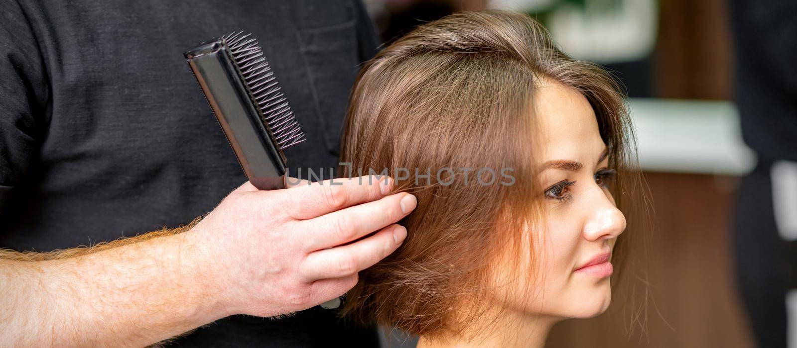 Male hairdresser works on the hairstyle of the young caucasian brunette woman at a hair salon. by okskukuruza