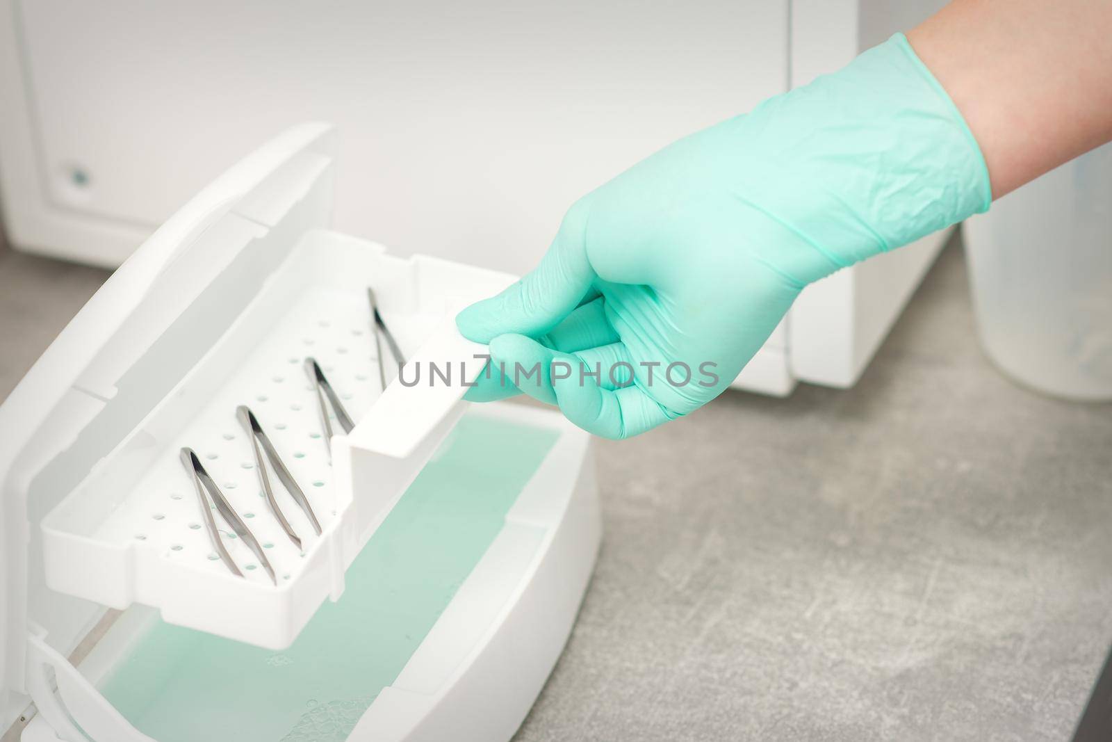 The beautician holds a tray of tweezers preparing them for disinfection with a special liquid and machine