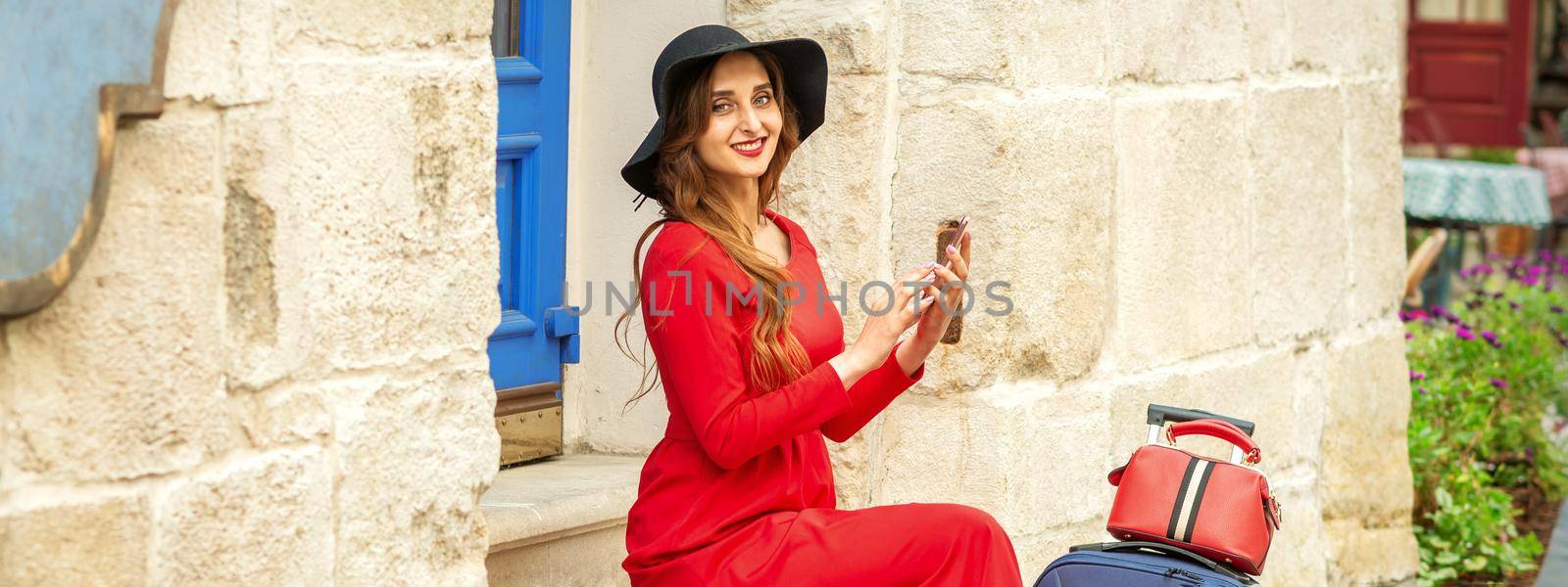 Portrait of tourist young caucasian woman in a red long dress and black hat with suitcase sitting on the stairs outdoors