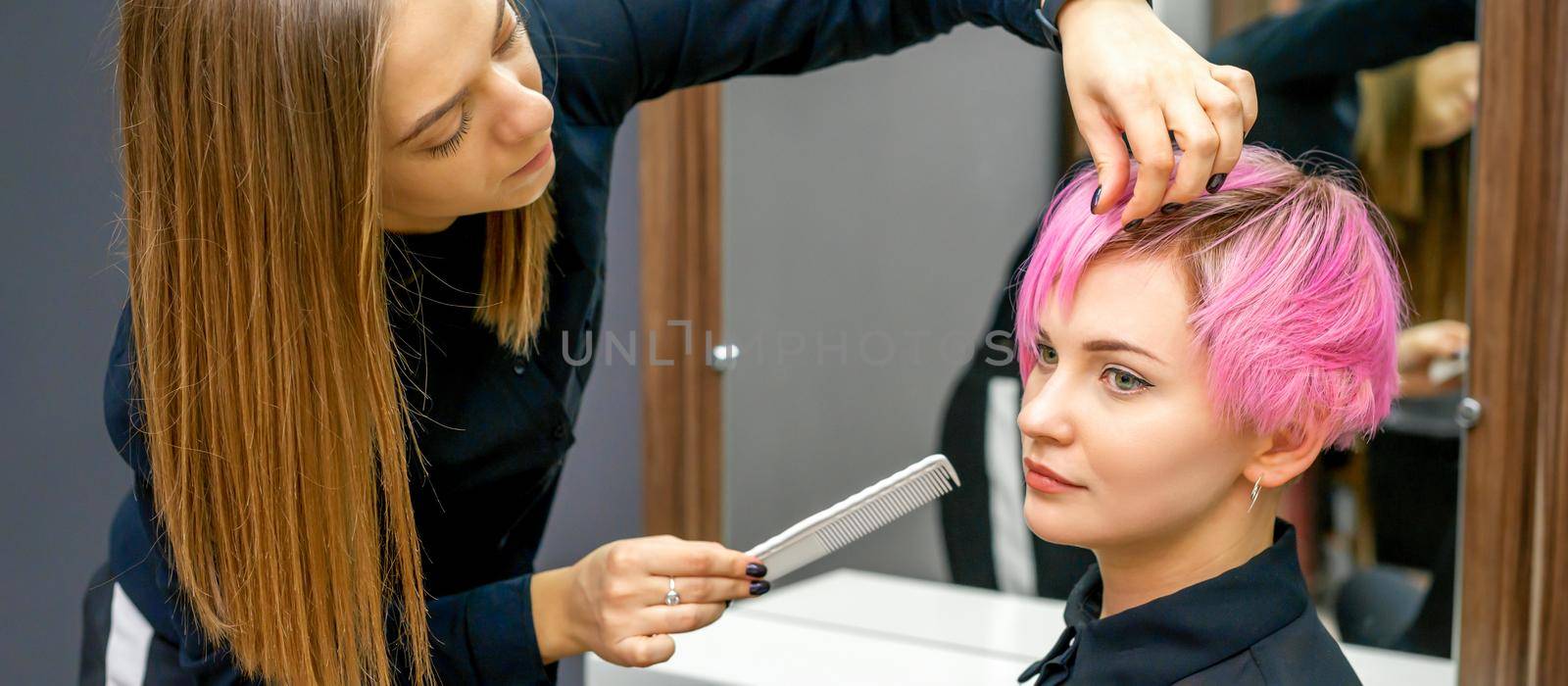 Female hairdresser styling short pink hair of the young white woman with hands and comb in a hair salon. by okskukuruza