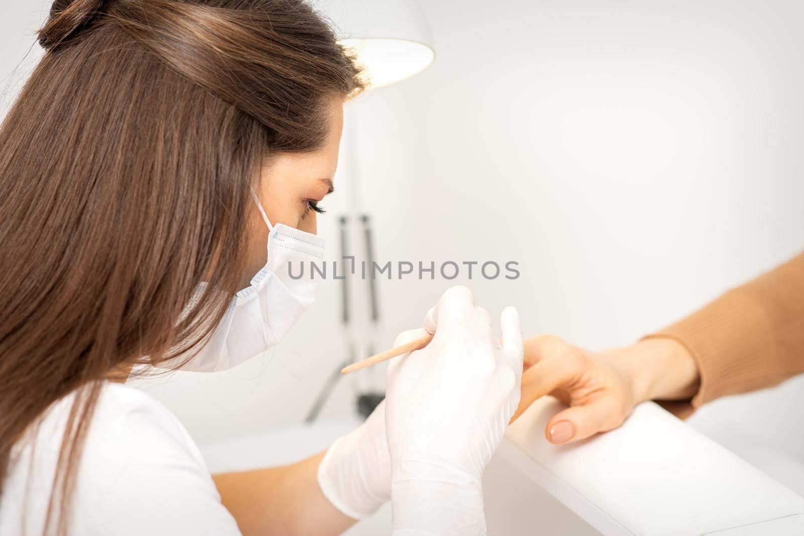 Manicure master in protective face mask and white gloves apply polish to female fingernails