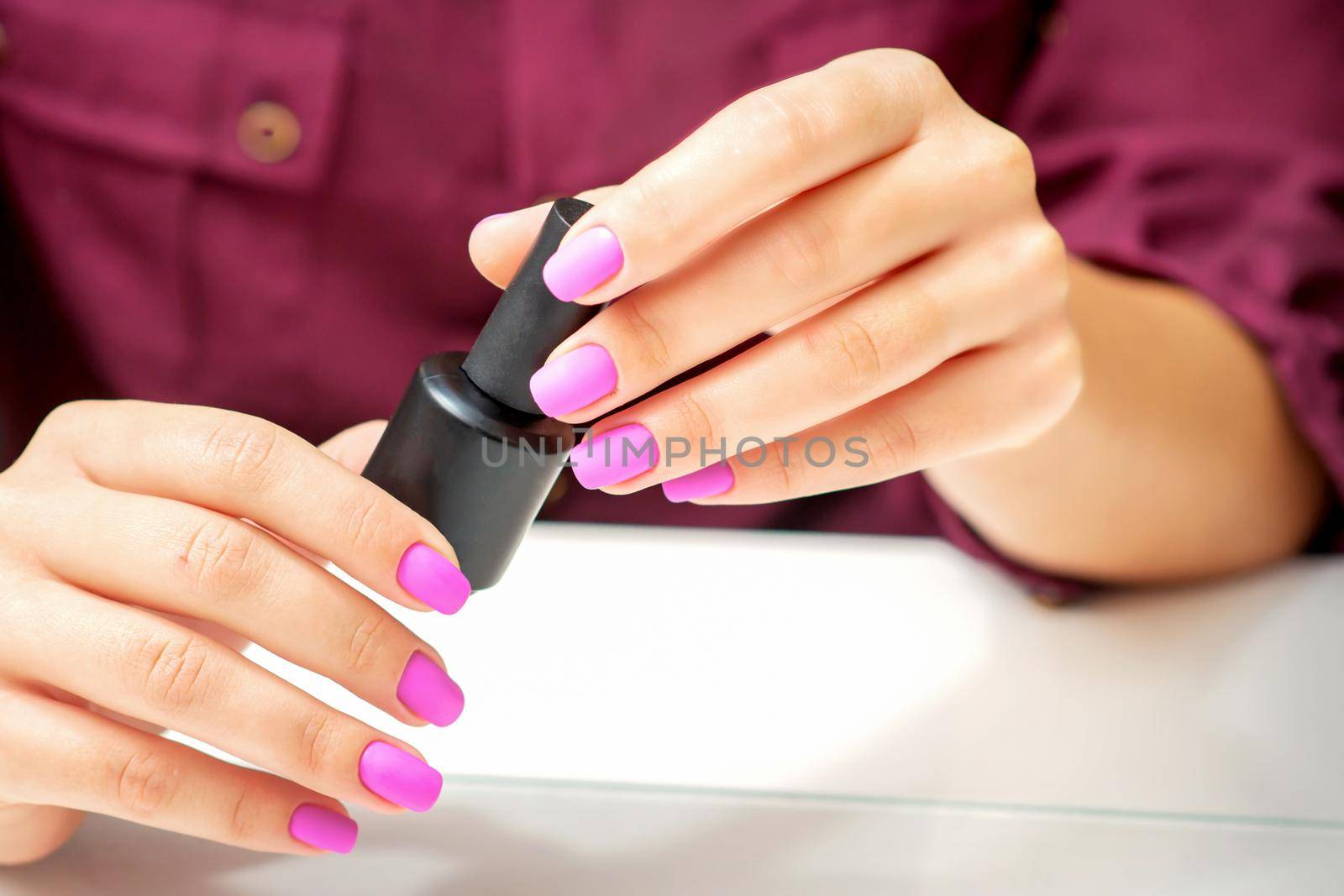 White woman holding nail polish black bottle with painted pink nails close up