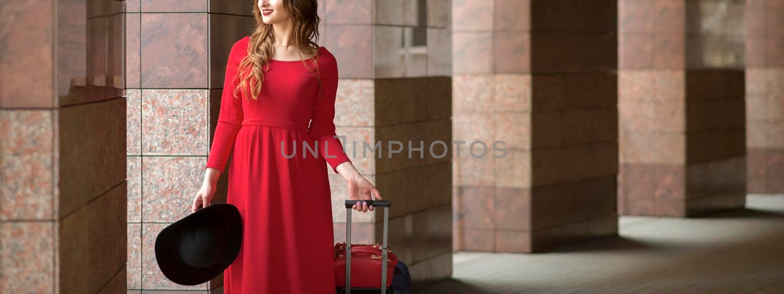 Beautiful young caucasian woman wearing red long dress with a suitcase standing in columns. by okskukuruza