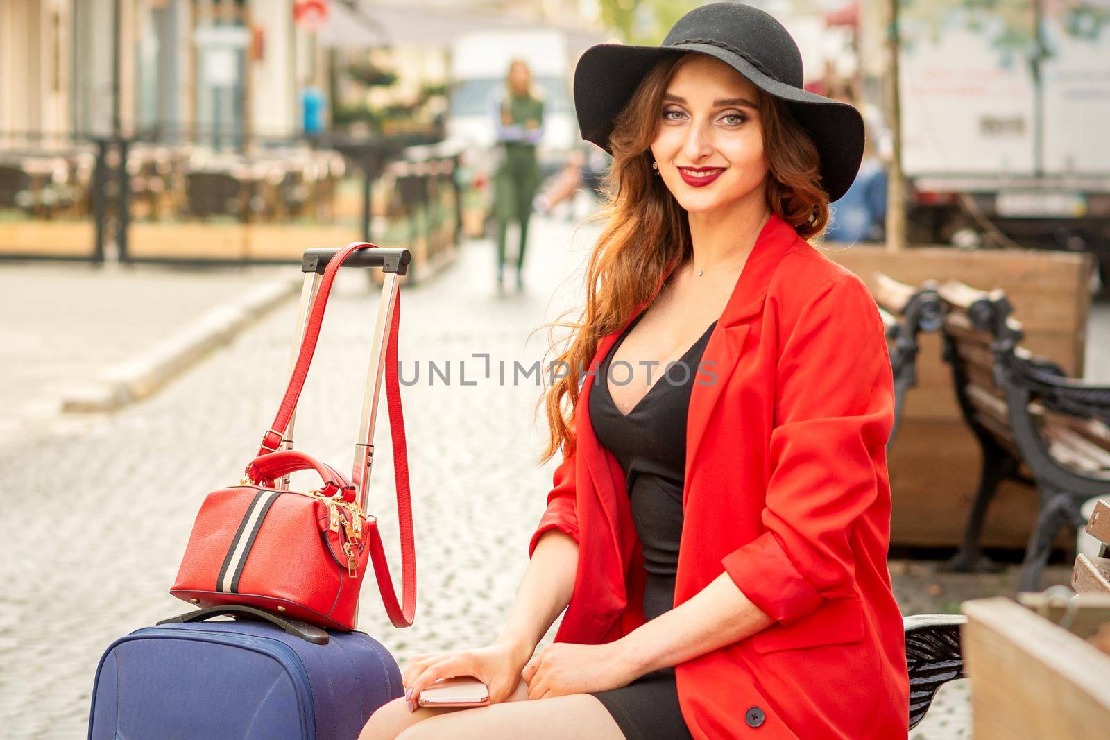 A young traveling white woman in the red jacket and black hat sitting with the suitcase on the city street