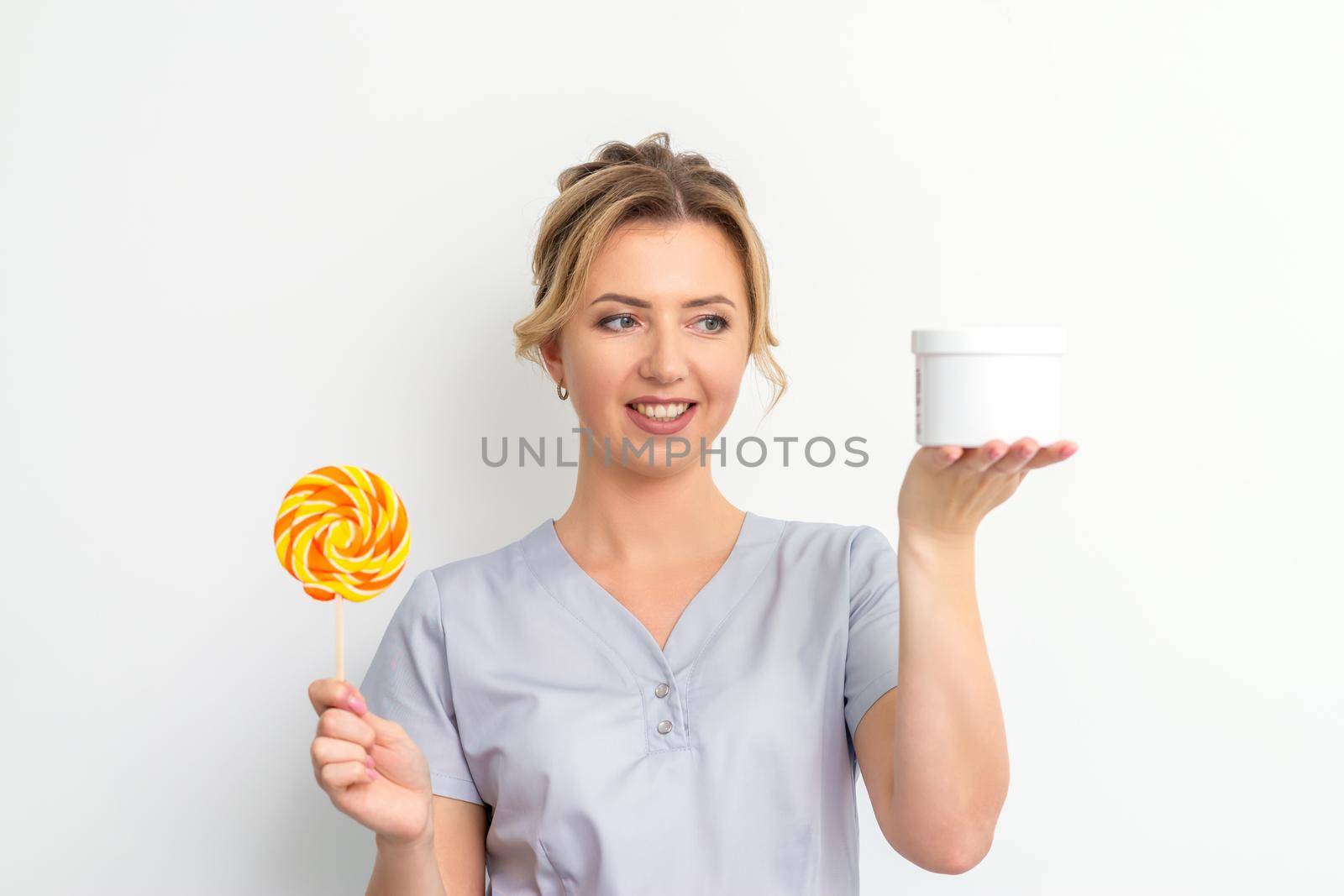 Beautician holding a jar of wax for waxing and candy on a stick smiling on a white background. Natural product for hair removal