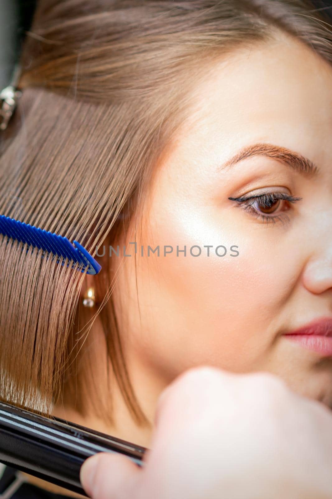 Hairdresser straightens female brown hair of medium length with a hair with an iron hair straightener and comb in a beauty salon. by okskukuruza