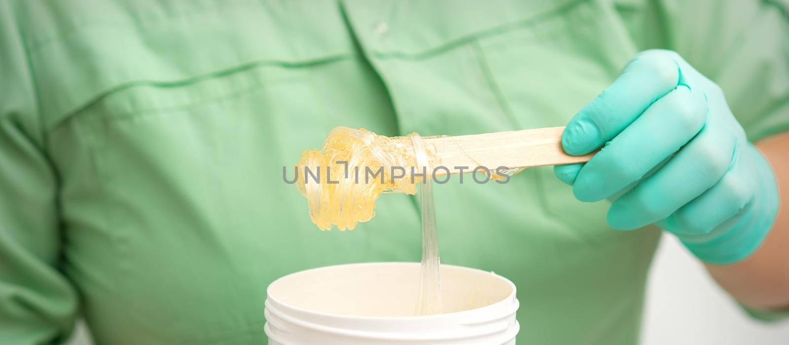 Liquid yellow sugar paste for depilation on a stick flows into the jar in hands of a beautician