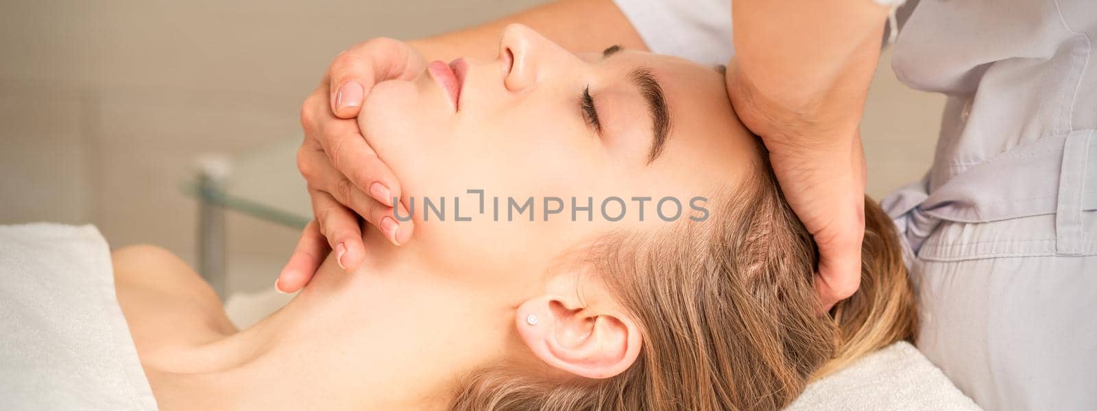 Cosmetologist hands doing facial massage on forehead and chin of young female at spa salon. by okskukuruza