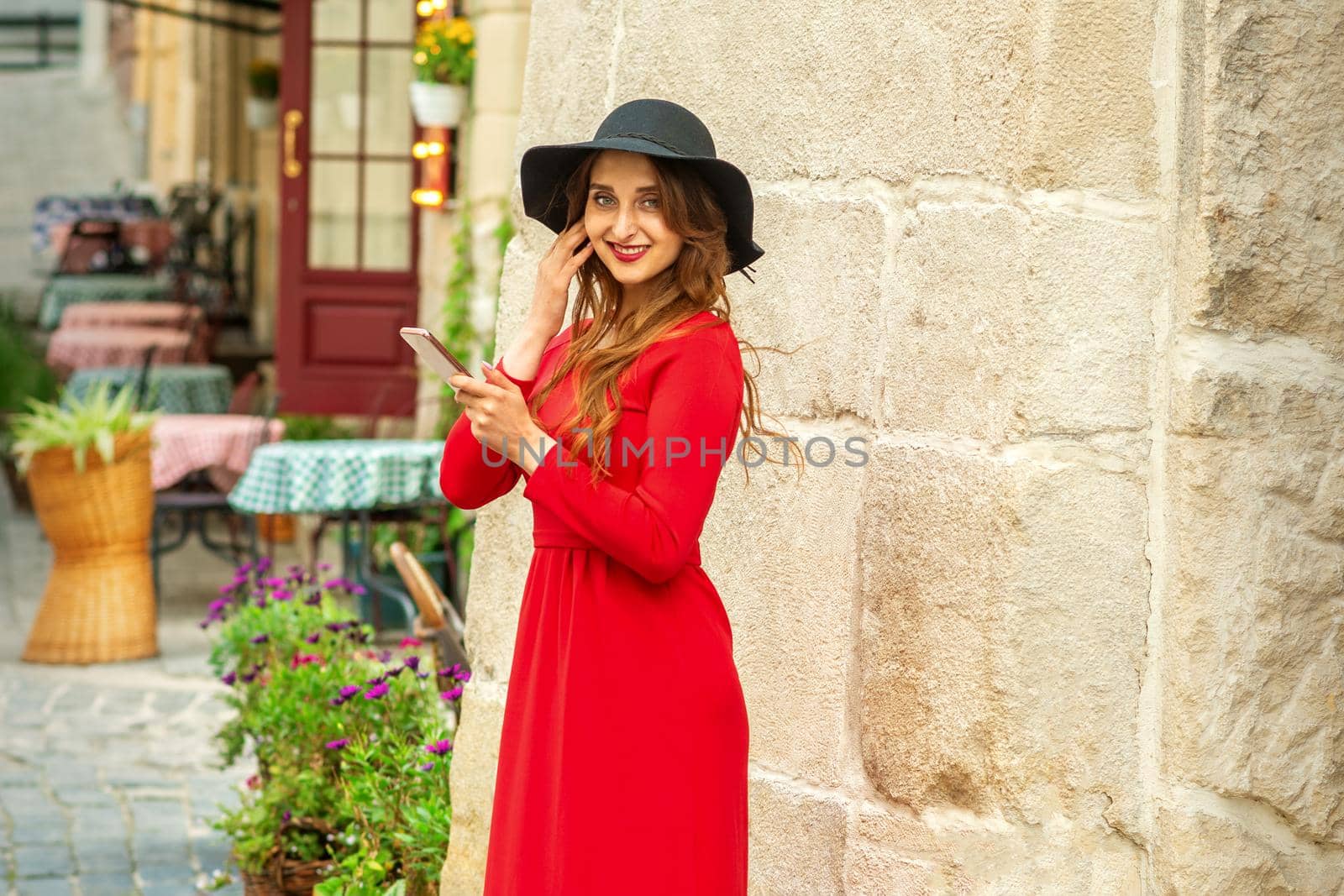 A young traveling white woman in the red jacket and black hat standing with a smartphone on the city street