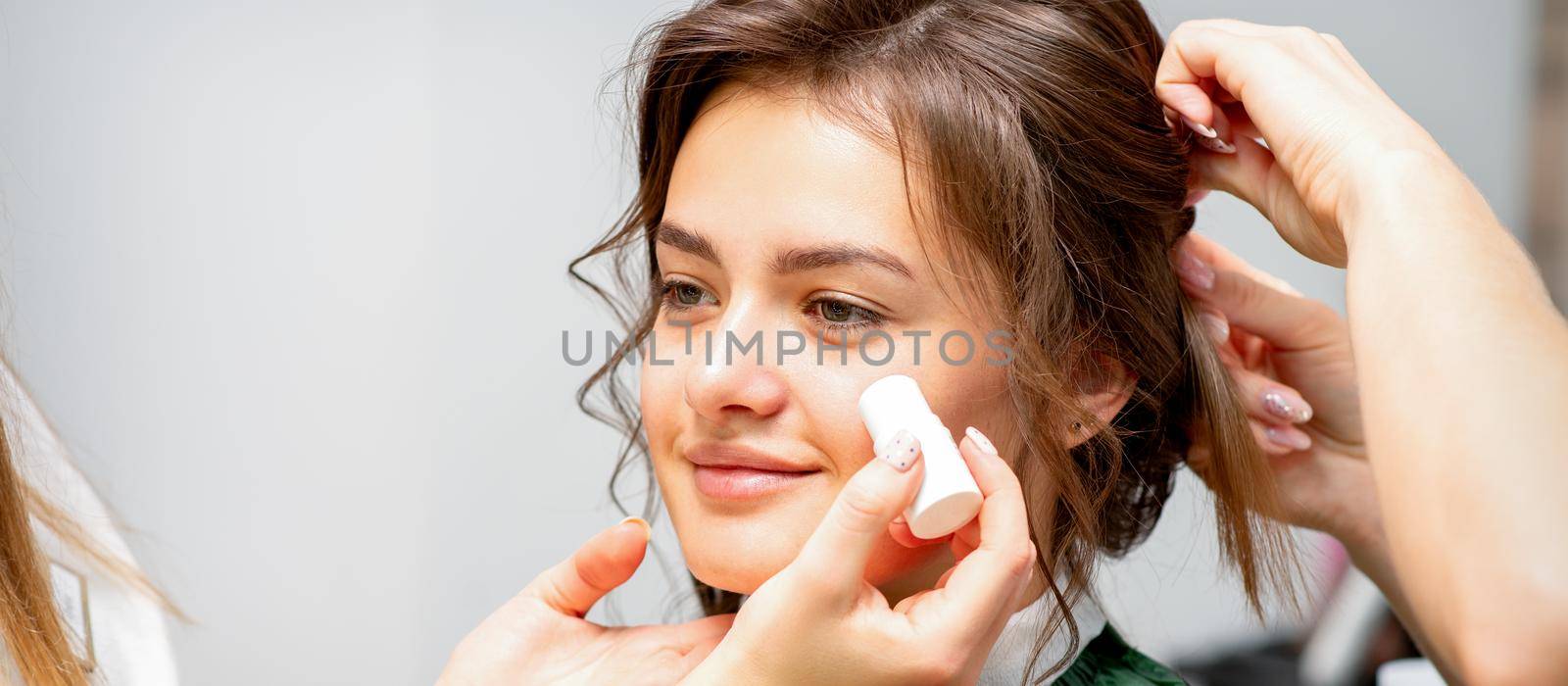 Makeup artist applying cream blush foundation tube on the cheek of the young caucasian woman in a beauty salon