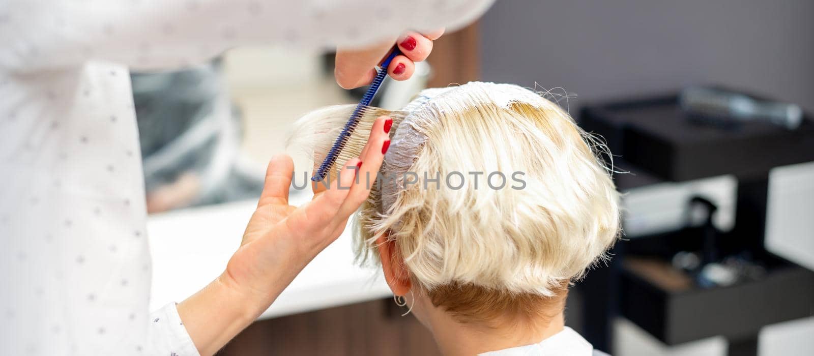 Female hairdresser styling short white hair of the young blonde woman with hands and comb in a hair salon