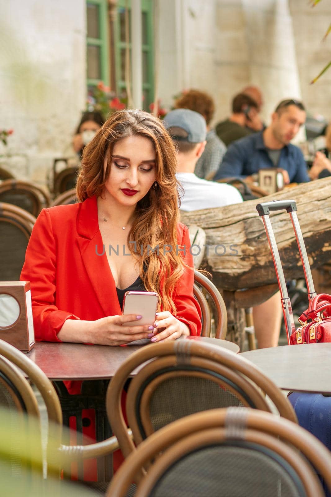 Tourist young caucasian white woman in a red jacket looking at smartphone sitting at the table in cafe outdoors