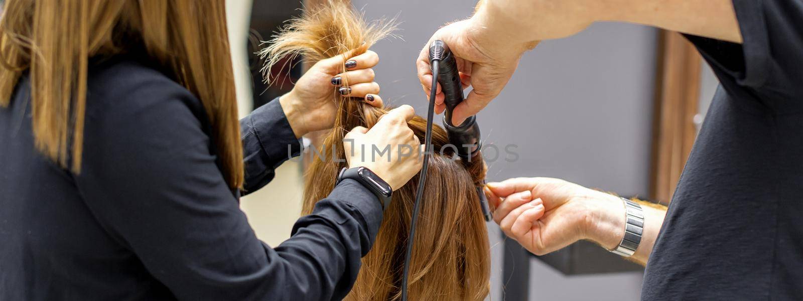 The rear view of two hairdressers are curling hair for a young woman with electric hair iron in a beauty salon. by okskukuruza