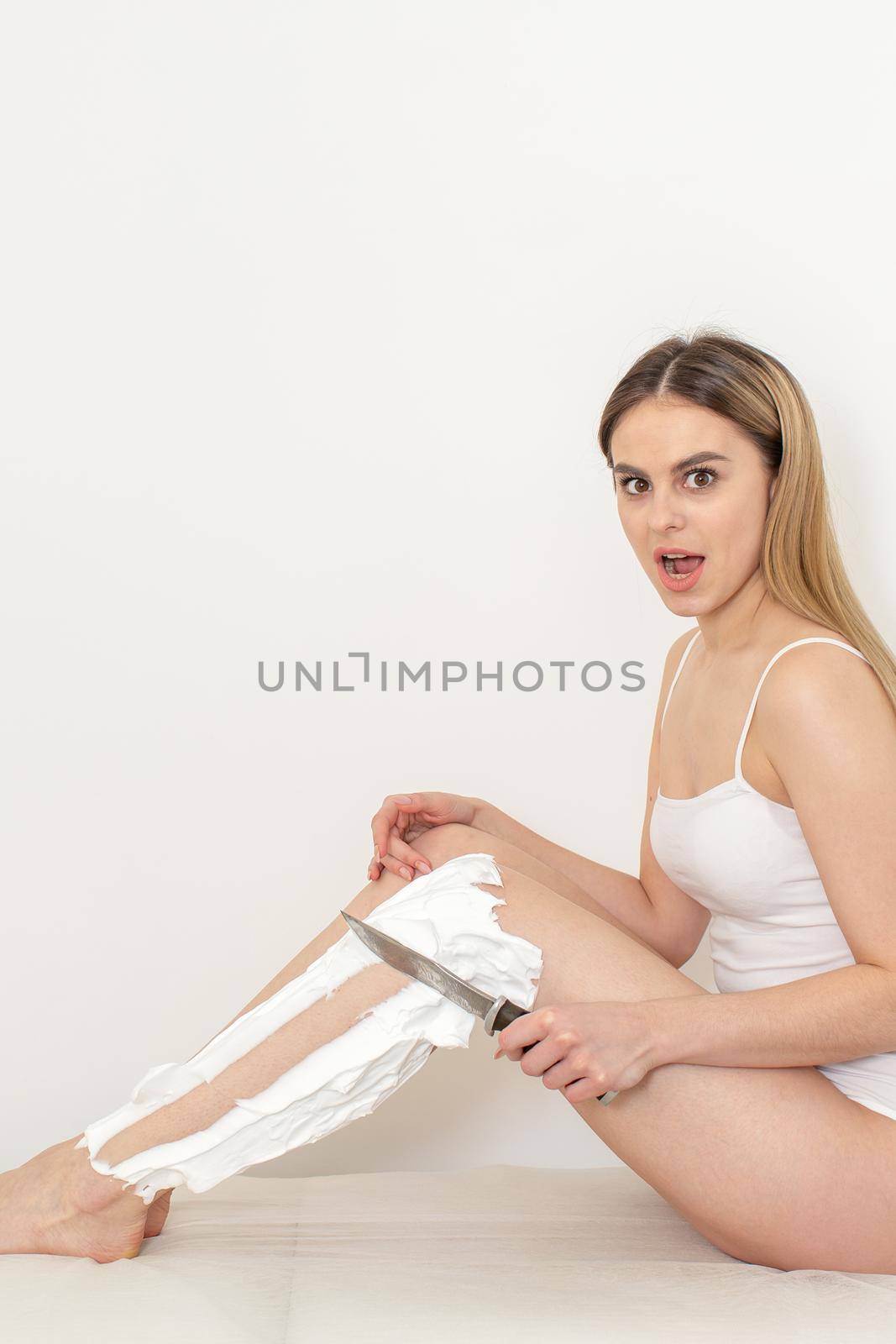 Beautiful caucasian woman shaving her legs with a knife with shaving foam on white background. Depilation and epilation concept. by okskukuruza