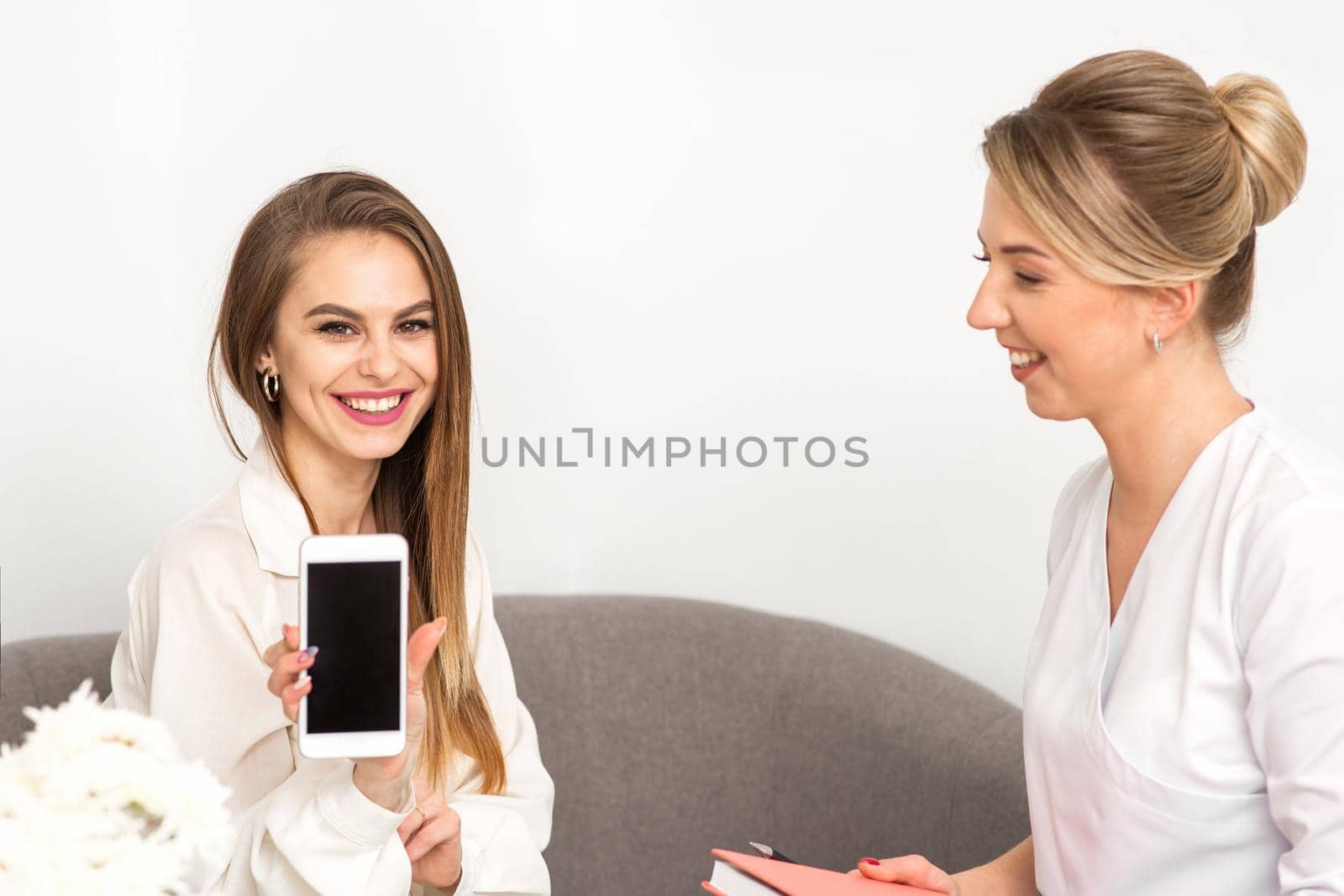 Two smiling female doctors wearing white medical gowns with empty smartphone screen in a medical office, mockup, copy space. by okskukuruza
